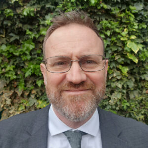 A photograph of Ed Finch, smiling, wearing a smart jacket and tie.