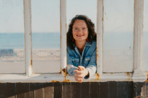 A smiling Celebrant Gitte (white woman, dark hair, light denim jacket) leaning through a white and rusty window on Brighton beach with the sea in the background.