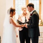 A bride and groom holding hands to say their vows with their humanist wedding celebrant standing between them holding a file.