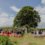 A large oak tree in the middle of a bean field, with straw bales arranged around it for the guests at a humanist wedding in Wiltshire