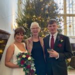 A couple and their Humanist wedding celebrant standing in front of a Christmas tree and smiling at the camera.