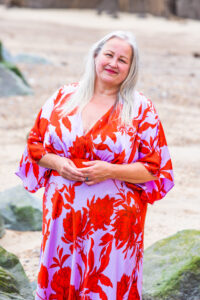 Picture of me on the beach, wearing a brightly patterned red and lilac dress, smiling warmly