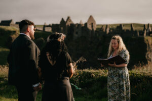 Laura, the celebrant, stands on the edge of a cliff on the Irish coast, with the historic ruins of Dunluce Castle in the background. A bride and groom, both dressed in black, are facing her.