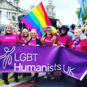 Laura and some colleagues stand outside Belfast City Hall on a march for same-sex marriage. The group are all wearing pink t-shirts with 'LGBT Humanists UK' on the front, and are holding a large purple banner bearing the LGBT Humanists UK and 'happy human' logo. One of the group is waving a huge Rainbow Pride flag, and all are beaming at the camera.