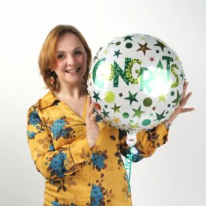 Lily holding a balloon that says 'Congrats!' and smiling at the camera in a yellow shirt.