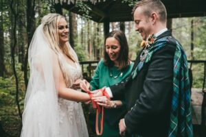 A celebrant in a green dress binds a bride and groom together with ceremonial ribbons. The wedding is taking place in wood, at Camp Katur in North Yorkshire.