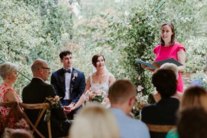 A wedding celebrant in a pink dress stands at a wedding. The bride and groom are seated beside her, watched by wedding guests. The wedding is happening in a leafy garden.