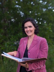 Raquel is smiling into the camera, while wearing a pink blazer and holding a folder in both hands. 