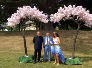 Simon with couple under a flower arch in front of trees.