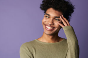 Happy trans gender person with a big smile, black hair and closed eyes with eye-shadow on a purple background.